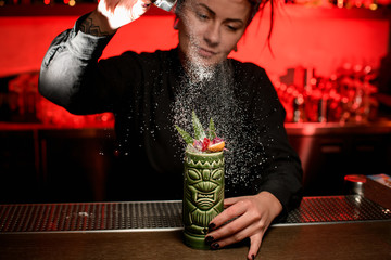 Pretty bartender girl serving alcoholic cocktail in the Tiki mug sprinkling on it with sugar powder