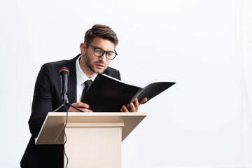 businessman in suit standing at podium tribune and holding folder during conference isolated on white