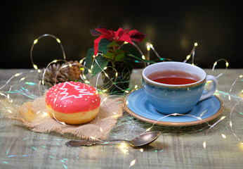 Sweet donut decorated for Christmas on a burlap with cup of tea, old silver teaspoon and Xmas flower. Romantic Christmas breakfast, dessert with magic lights of garland. Poinsettia plant.