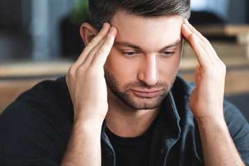 handsome man with headache touching head and looking down in apartment