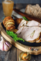 A piece of baked stuffed meat with potatoes and vegetables in a frying pan and a wooden Board