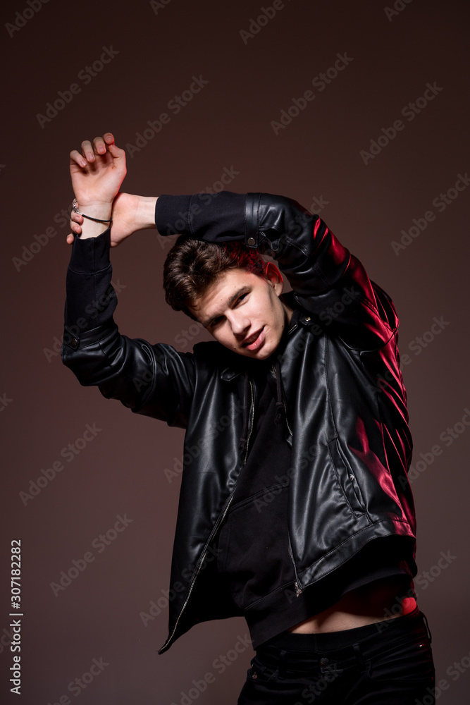 Wall mural Attractive young guy posing in studio. Young caucasian guy model in black clothes posing in the studio on a dark burgundy background with red backlight. Man in a leather jacket