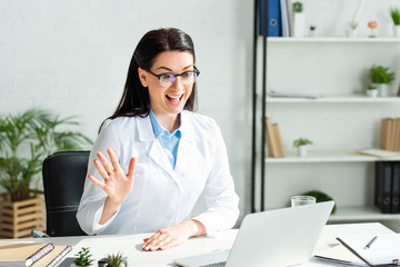 excited doctor waving and having online consultation with patient on laptop in clinic office