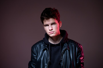 Attractive young guy posing in studio. Young caucasian guy model in black clothes posing in the studio on a dark burgundy background with red backlight. Man in a leather jacket
