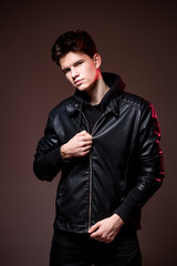 Attractive young guy posing in studio. Young caucasian guy model in black clothes posing in the studio on a dark burgundy background with red backlight. Man in a leather jacket