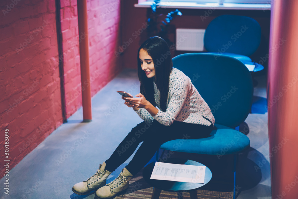 Aufkleber smiling hipster girl blogging in networks and reading news via smartphone during free time in cowork