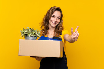 Naklejka premium Young blonde girl making a move while picking up a box full of things smiling and showing victory sign