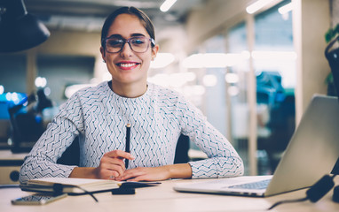 Portrait of cheerful businesswoman satisfied with occupation sitting at desktop in office with...
