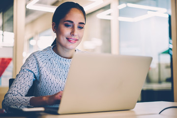Positive female manager satisfied with working process browsing information for project on laptop computer, smiling woman watching video from networks on netbook spending time at desktop in office