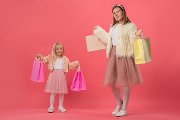 smiling daughter and mother holding shopping bags on pink background
