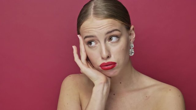 A bored young woman with red lipstick and earrings is looking to the side isolated over red background