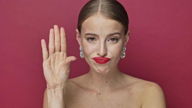 A bored young woman with red lipstick and earrings is closing her ears with her fingers isolated over red background