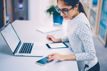 Brunette businesswoman working in office checking notification on smartphone synchronizing netbook...