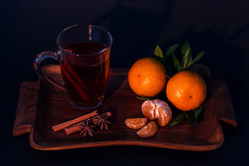 Still life. On a wooden tray is a glass cup with tea and cinnamon, ripe tangerines. Black background. Close-up.