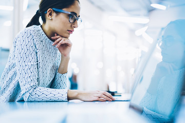 Concentrated woman employee in eyewear for vision protection reading information from laptop...