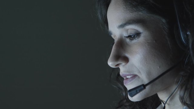 Female teleworker working at night. Close-up view of focused young female call center operator in headset talking and working in dark office. Client support concept