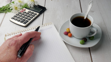 Desktop with cup of coffee, calculator, notepad, pen and white flowers in the background. Accounting business concept.  Female hand holds a pen.