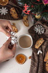 Broken chocolate chip cookies in the hands of a man over a cup of coffee with milk on a table with traditional Italian biscotti cookies with on a gray background with slices of dried oranges