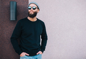 City portrait of handsome hipster guy with beard wearing black blank hoody or sweatshirt and hat...