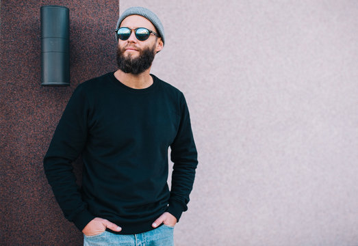City portrait of handsome hipster guy with beard wearing black blank hoody or sweatshirt and hat with space for your logo or design. Mockup for print