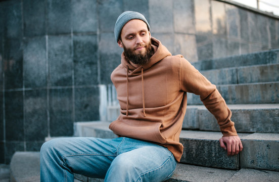 City portrait of handsome hipster guy with beard wearing brown blank hoodie or hoody and hat with space for your logo or design. Mockup for print
