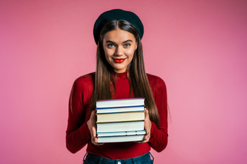 Pretty student on pink background in studio holds stack of university books from library. Girl in hat smiles, she is happy to graduate.