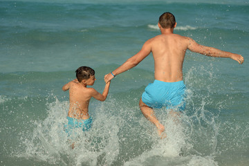 Cute European boy is splashing in the sea with his dad. They are having fun during their holidays on the seacoast.