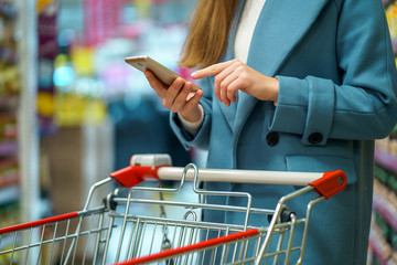 Woman buyer with cart in the shop aisle with grocery list on smartphone during shopping food