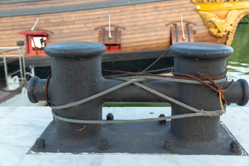 Fototapeta na wymiar Cast iron, black, with signs of corrosion mooring device with a wound steel cable is fixed to the concrete pier studs and nuts on the background of the side of the wooden ship.