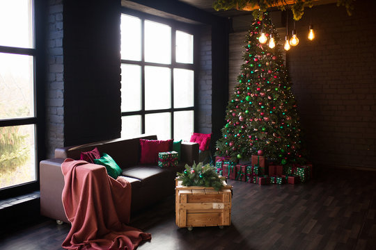 Dark loft interior. Christmas tree with a bunch of boxes with gifts. A beautiful green leather sofa on the background of a gray brick wall with panoramic window.