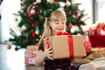 Happy girl sitting next to christmas tree holding present