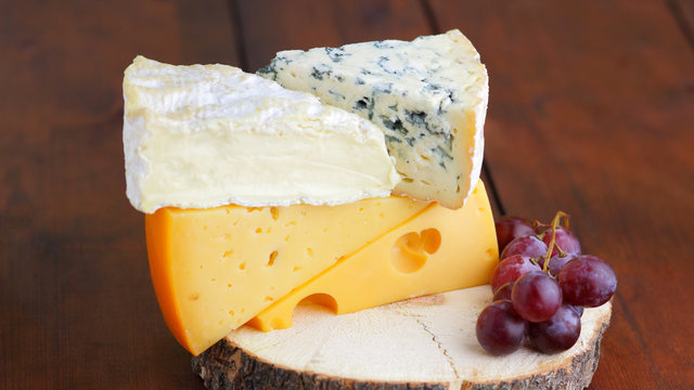 Camembert, brie, hard cheese and blue cheese on wooden board. Slices of various cheeses and grapes on blurred background. Dorblu and soft cheese on wooden boards. Widescreen