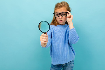 curious european girl in glasses looks through a magnifying glass on a light blue background