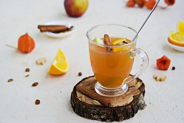 Apple cider with spices. Alcoholic or non-alcoholic hot drink made from fresh unfiltered apple juice with spices, apple slices and orange in a glass mug on a light concrete background.