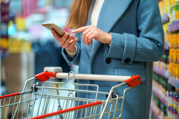 Woman buyer with cart in the store aisle with grocery list on smartphone during shopping food
