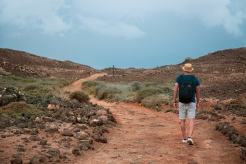 Hombre con sombrero y mochila andando por camino de tierra sobre montaña y rocas con cielo azul...