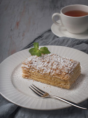 Tasty dessert piece of cake with white powder sugar with a leaf of mint close-up on a white plate on the background of a tea cup on a gray textured concrete background