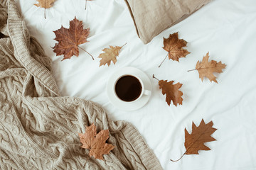 Cup of coffee with milk, dry leaves on bed with white linen, pillow, blanket. Flat lay, top view. Minimal fall autumn morning breakfast in bed composition.