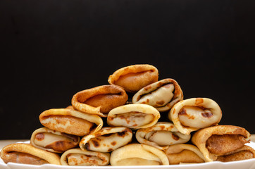Many homemade thin pancakes with filling stacked pyramid on a white plate against a black wall, copy space