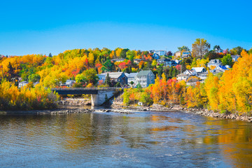 Fototapeta premium Jesienny krajobraz Widok wsi nad brzegiem jeziora z kolorowych drzew, rzeki i mostu na tle błękitnego nieba odbite w wodzie