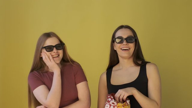Two girls in glasses are looking a film in a cinema, eating popcorn and laughing