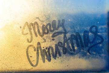 Merry Christmas written on wet window glass at sunset light. Beautiful blue and yellow background of drops and splashes, concept for New Year and Christmas