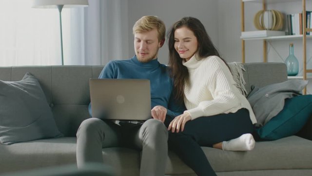 
Cute Young Couple Use Laptop Computer, Have Fun, Laughs, while Sitting on the Couch in the Cozy Apartment. Couple Shopping on Internet, Using Social Media, Watching Videos and Streaming Content 