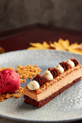 Slice of layered cake with dark chocolate biscuit, milk chocolate mousse, caramelized nuts, decorated with whipped cream and garnished with ball of berries ice cream.