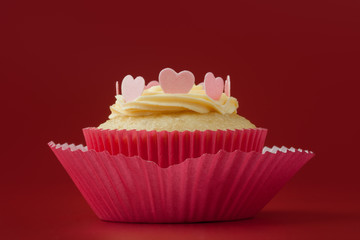 Baked vanilla cupcake with icing sugar and pink eatable hearts in decorative paper mold, red background