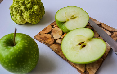 Tasty food in green. Two apples: one is whole, the other cut into two parts. Broccoli in a strange pyramid shape. Knife and board for cutting made of wood.