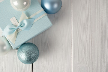 Gift box of pale blue color with blue and white bow and Christmas balls of white and blue color on a wooden white table with a white brick background.
