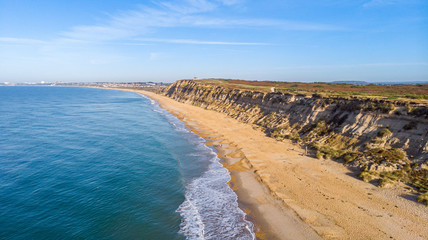 An aerial view of a majestic seacoast hill with huge cliff, sandy beach, groyne (breakwater) and crystal blue water under a beautiful blue sky and some white clouds