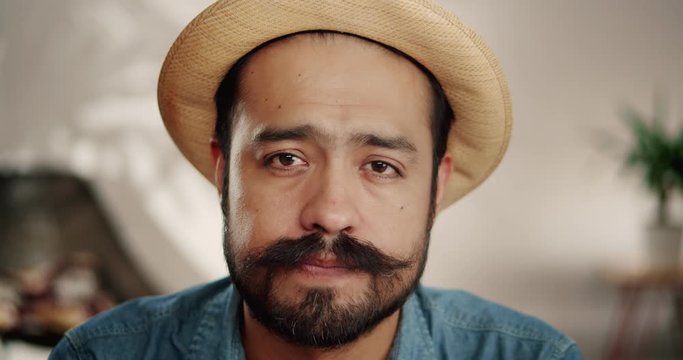 Close up of Hispanic adult man in hat with mustache feeling sadness looking to camera, sorrowful handsome naive bearded male with brown eyes dressed in jeans shirt indoor, disappointed perplexity