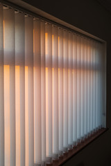 White vertical slat blinds hanging in front of a window as the sun is setting turning the light golden. The slats have sealed glued pockets and no cords at the bottom. Soft shallow focus